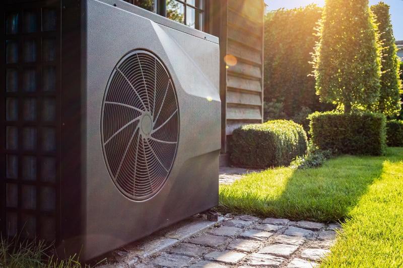 Transitioning Your Heat Pump Use Between Seasons. Close-up of black full inverter heat pump outside in the garden, near wooden pool house on a sunny day.